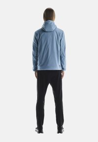 Veste à capuche bleu clair avec une texture lisse et des manches longues, associée à un pantalon noir. La tenue présente une coupe décontractée et peu de détails.