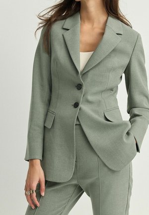 Kobieta w dopasowanej, szałwiowozielonej marynarce z dwoma czarnymi guzikami, dopasowanych spodniach, białym topie pod spodem i pierścionkach na lewej ręce.