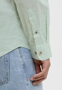Camasa din in verde deschis cu maneci lungi, dotată cu manșete cu nasturi și o croială lejeră, asortată cu blugi cu talie înaltă.