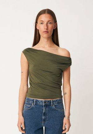 Kvinde med langt brunt hår iført olivengrøn top med ét skulder og blå jeans, stående mod en ensartet lys baggrund.