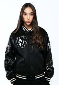 Chaqueta varsity negra con mangas de satén, cuello con rayas blancas y varios logotipos bordados en el pecho y las mangas.