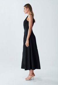 Černé bezrukávové šaty s přiléhavým korzetem a volnou sukní až po kotníky. Modelka má na sobě bílé sandály na podpatku. Hladká textura látky.