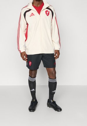 Mężczyzna ubrany w białą i czerwoną kurtkę Liverpool FC, czarne spodenki z logo Liverpool, czarne, sięgające kolan skarpety Adidas oraz czarne korki piłkarskie.
