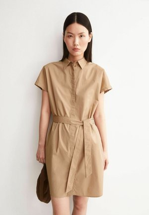 Frau mit glattem dunklem Haar trägt ein kurzärmeliges beigefarbenes Hemdblusenkleid mit Taillengürtel und hält einen braunen Hut vor weißem Hintergrund.