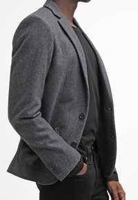 Šedý vlněný blazer se strukturovaným střihem, s klopami, dvěma knoflíky a bočními kapsami, nošený přes �černou košili a kalhoty.