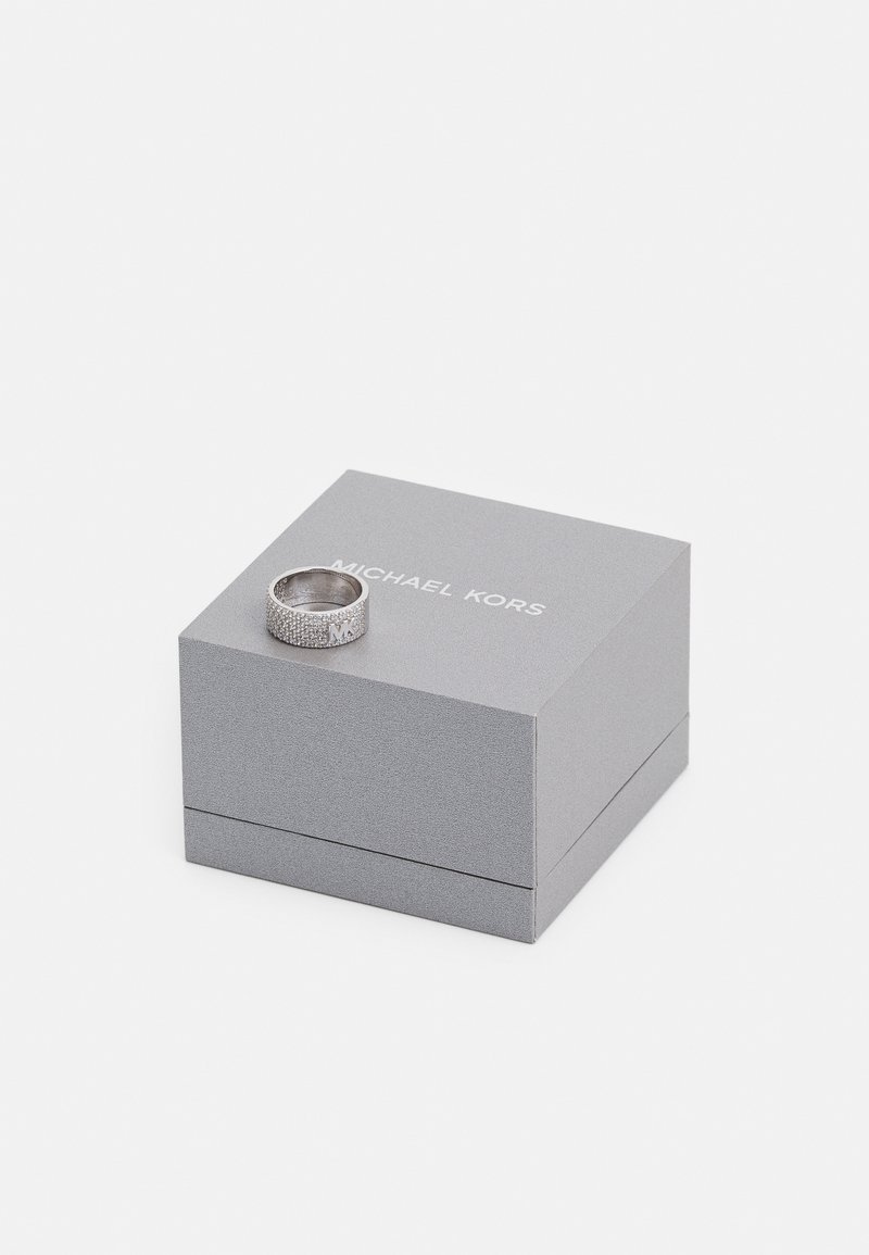 Anillo de plata con banda texturizada y detalle del logotipo, colocado sobre una caja rectangular gris etiquetada "MICHAEL KORS". La caja tiene un acabado suave.