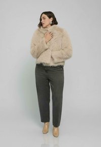 Kurtka z futra ekologicznego w beżowym kolorze, z wysokim kołnierzem i szerokimi rękawami, w zestawieniu z ciemnymi dżinsami i beżowymi botkami za kostkę, na jasnoszarym tle.