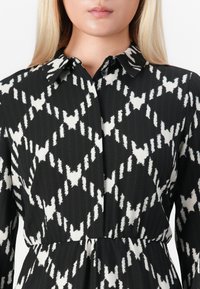 Femme portant une robe boutonnée à motifs géométriques noir et blanc, avec col et manches longues, cintrée à la taille.