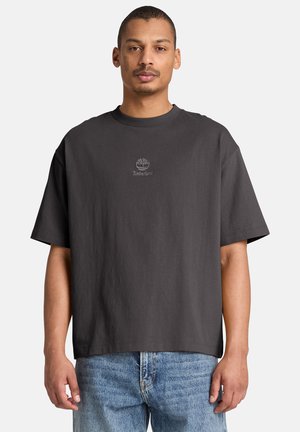 Man draagt een donkergrijs oversized Timberland T-shirt en lichtblauwe spijkerbroek, staand tegen een eenvoudige lichte achtergrond.