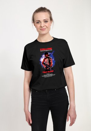 Kvinde iført en sort T-shirt med en farverig "First Blood" filmplakat med en muskuløs mand, der holder en pistol, og rød tekst.