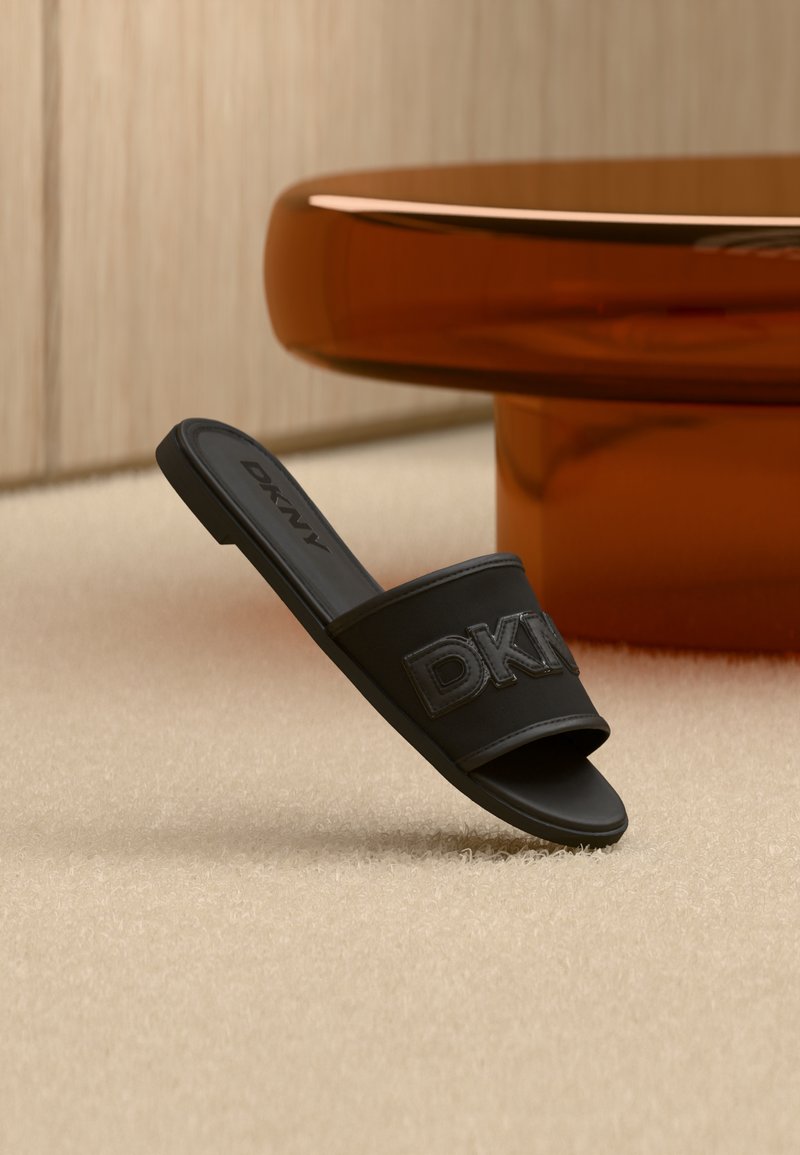 Μαύρο σανδάλι DKNY slide πάνω σε μπεζ χαλί δίπλα σε στρογγυλή βάση τραπεζιού κεχριμπαρένιου χρώματος σε δωμάτιο με ουδέτερους τόνους.