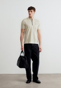 Νεαρός άνδρας στέκεται, φορώντας μπλούζα με μπεζ κοντά μανίκια και φερμουάρ, μαύρο παντελόνι, μαύρα παπούτσια, κρατώντας δίπλα του μια μαύρη τσάντα duffle.