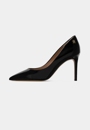 Sorte patentlæder højhælede pumps med spids tå, en slank stiletto hæl og et lille guld dekorativt logo nær hælen.