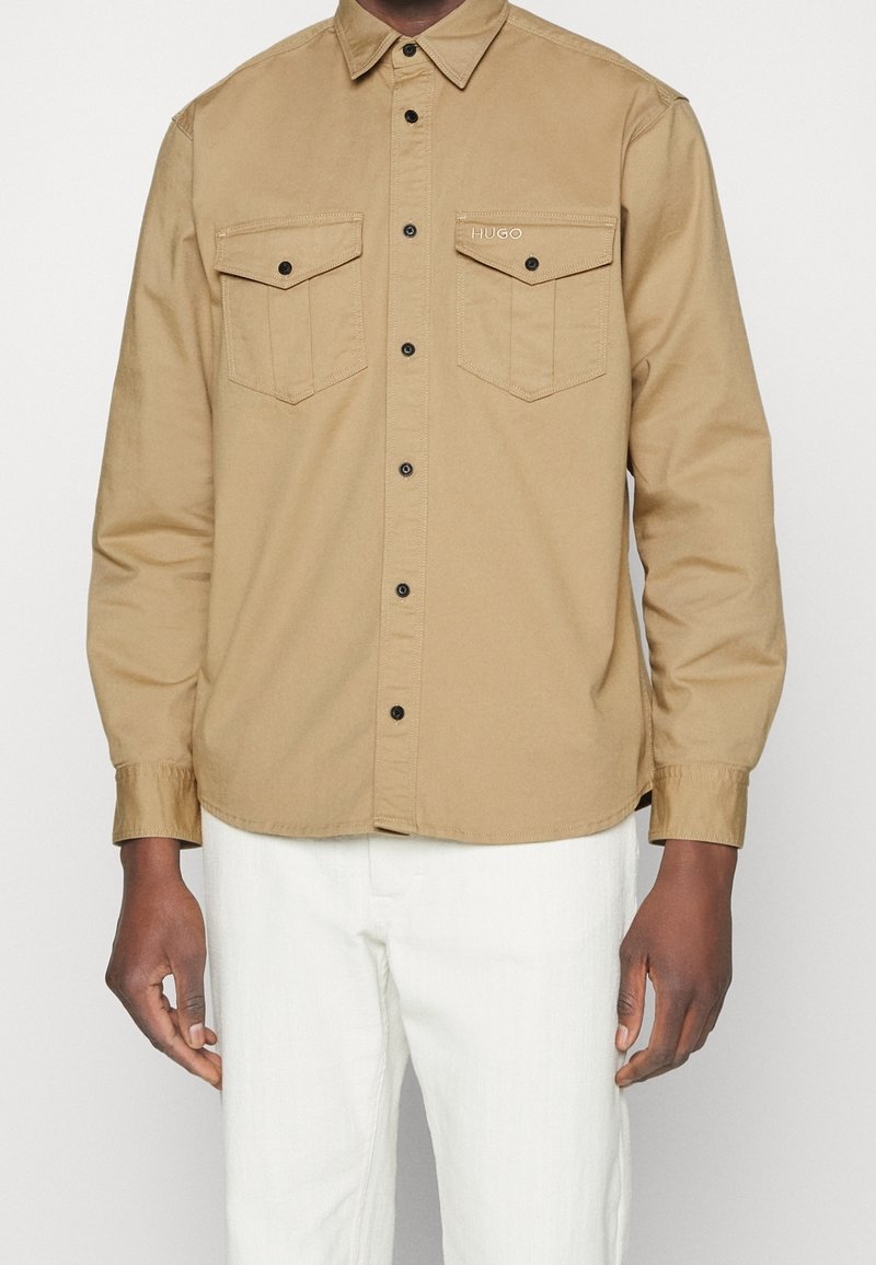 Hombre vestido con una camisa beige de botones con dos bolsillos en el pecho, botones negros y pantalones blancos frente a un fondo liso.