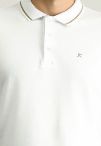 Biała koszulka polo wykonana z miękkiej tkaniny, z ryflowanym kołnierzykiem z złotym akcentem oraz małym logo wyszytym na piersi.