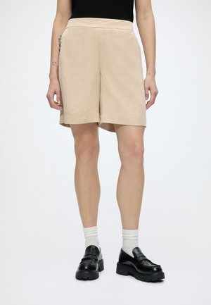 Személy bézs térd hosszúságú rövidnadrágot, fekete vastag talpú cipőt, fehér zoknit és fekete felsőt visel, fehér háttér előtt áll.