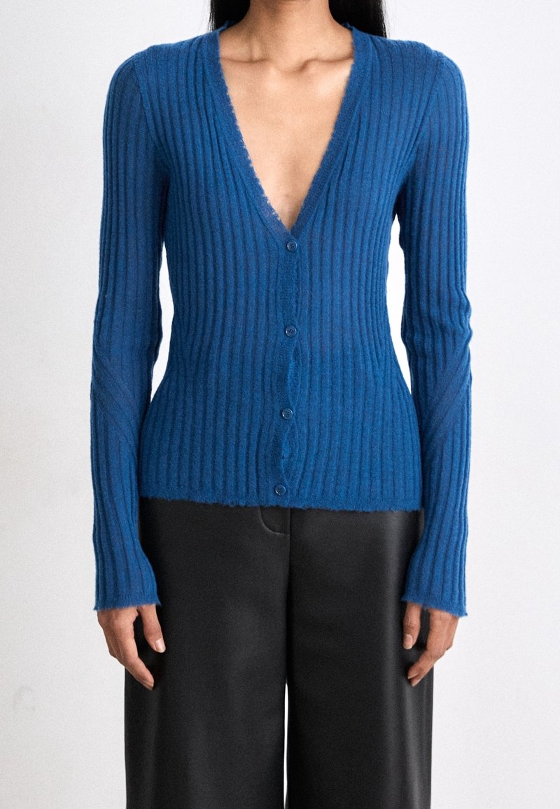 Femme portant un cardigan bleu côtelé ajusté à manches longues avec des boutons et un pantalon noir à jambes larges sur un fond uni.