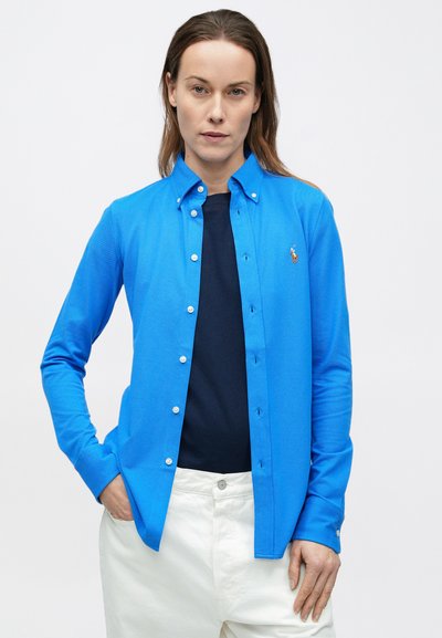 Modrá košeľa s dlhým rukávom na gombíky z textúrovaného materiálu s bielymi gombíkmi a malým vyšívaným logom na hrudi, kombinovaná s námorníckym vrchným dielom a bielymi nohavicami.