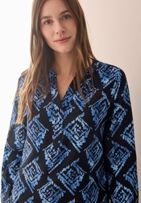 Blusa a maniche lunghe nera con un motivo geometrico blu; presenta un collo a V e una texture morbida. Il design include forme di diamante e tonalità di blu diverse.