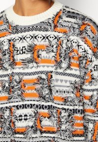 Strickpullover mit strukturiertem Muster aus orangefarbenen, grauen und weißen Streifen. Das Design umfasst ausgefranste Akzente und geometrische Formen.