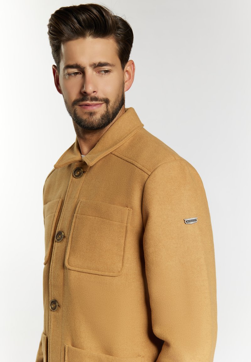 Veste en mélange de laine tan avec un col, deux grandes poches avant et une étiquette logo sur la manche gauche. Texture douce et design ajusté.