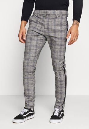 Pantalons à carreaux gris avec une coupe ajustée, présentant une bande noire sur le côté. Fabriqués en tissu à texture lisse. Associés à des baskets noires.