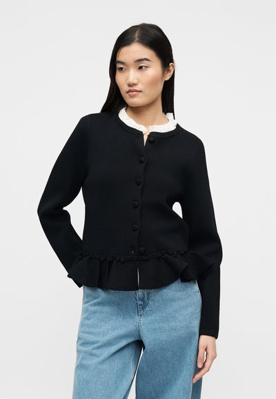 Γυναίκα που φοράει μαύρο πλεκτό ζακέτα με κουμπιά και βολάν στο τελείωμα πάνω από λευκό πουκάμισο, συνδυασμένο με ανοιχτό μπλε τζιν, στέκεται μπροστά σε απλό φόντο.