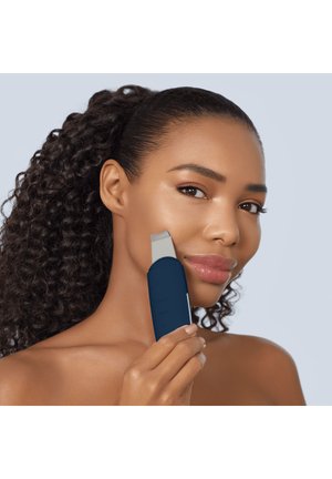 Femme aux cheveux bouclés utilisant un appareil de nettoyage du visage bleu foncé sur sa joue, sur un fond gris clair.