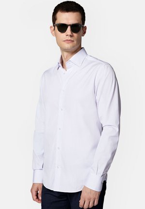 Biała koszula z guzikami, o usztywnionym kołnierzu, z długimi rękawami i delikatnym wzorem w pionowe paski, zestawiona z czarnymi okularami przeciwsłonecznymi.