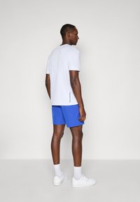 Mann som står med siden mot, iført hvit kortermet skjorte, blå shorts, hvite sokker og hvite joggesko mot en ensfarget bakgrunn.