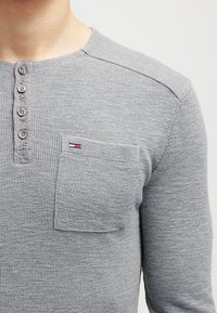 Šedé dlouhé tričko henley s texturovaným pleteným vzorem, s pěti knoflíčky, a jednou náprsní kapsou; obsahuje malý detail loga.
