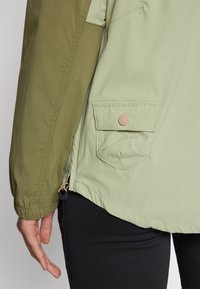 Lys grønn og oliven jakke med en sidelomme med en beige knapp, elastiske ermer og justerbar nedre kant med en snordetalj.