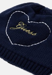 Tamsiai mėlyna megzta beanie kepurė su raukšlėta juosta, viduryje esantis baltos širdies kontūras ir auksinė siuvinėjimo "Guess" tekstas. Minkšta tekstūra.