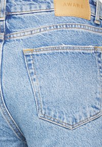 Jeans de denim azul claro com um bolso traseiro com costura amarela e uma etiqueta de couro com a palavra "AWARE" estampada.