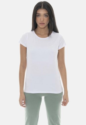 Femme aux longs cheveux foncés portant un t-shirt blanc uni à manches courtes et un pantalon vert clair, debout devant un fond clair.