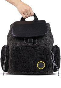 Mochila negra y gris con un exterior texturizado y esponjoso, solapa de cuero, dos bolsillos frontales y un logotipo circular con acentos amarillos.