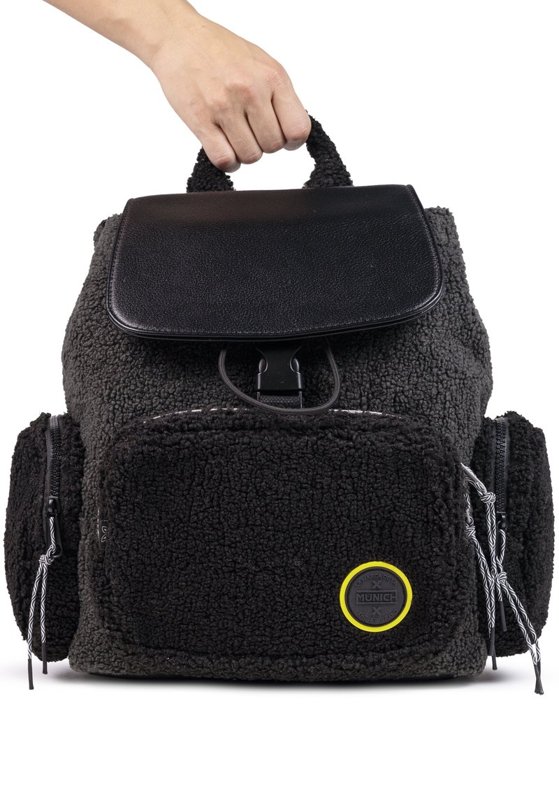 Mochila negra y gris con un exterior texturizado y esponjoso, solapa de cuero, dos bolsillos frontales y un logotipo circular con acentos amarillos.