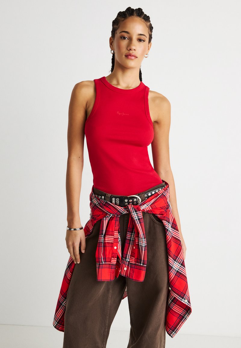 Νέα γυναίκα με πλεξούδες στα μαλλιά, φορώντας κόκκινο αμάνικο τοπ, καφέ παντελόνι και κόκκινο καρό πουκάμισο δεμένο γύρω από τη μέση, σε λευκό φόντο.