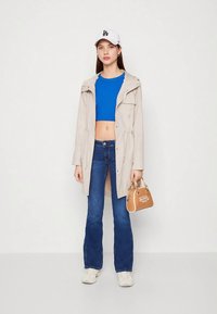 Šviesiai smėlio spalvos tranšėjos paltas, mėlynas trumpas viršus, platėjantys mėlyni džinsai, balti sportbačiai ir ruda rankinė su logotipo detalėmis, nufotografuoti neutraliame fone.