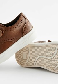 Zapatillas de cuero marrón con detalles perforados, suelas de goma blanca y partes inferiores con textura. Presentan un diseño elegante y redondeado.