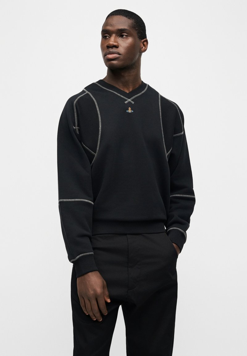 Giovane uomo con maglione nero con dettagli di cuciture grigie e piccolo emblema, mani nelle tasche, che guarda di lato contro uno sfondo bianco neutro.