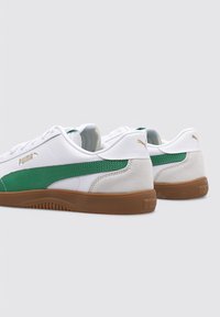 Zapatillas deportivas blancas con acentos verdes, detalles de ante y suelas de goma marrón. Presenta un diseño texturizado y un logo dorado en el lateral.