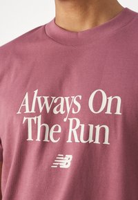 Close-up van een persoon die een mauve t-shirt draagt met witte tekst "Altijd op de vlucht" en een klein New Balance-logo eronder.