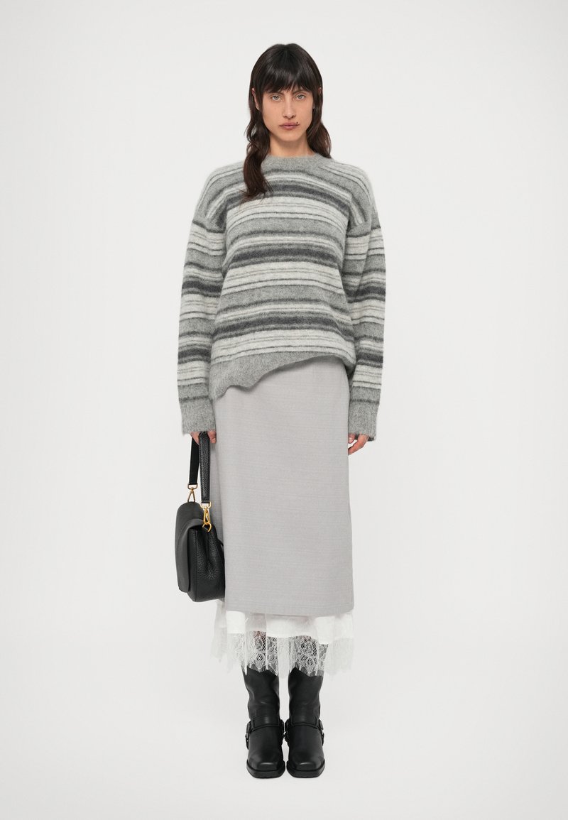 Sivi prugasti vuneni džemper opuštenog kroja, uparen sa svijetlosivom midi suknjom i crnim čizmama koje sežu do koljena. U ruci se drži crna torba.