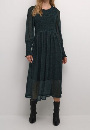 Nainen pukeutuneena tummanvihreään, pitkähihainen midi-mittainen mekko, jossa on kuminauhoitettu yläosa ja hihansuut, yhdistettynä mustiin midisäärisiin korkkareihin.