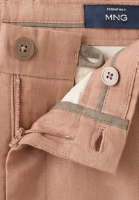 Gros plan sur la ceinture d'un pantalon rose montrant deux boutons, une fermeture éclair, une boutonnière et une étiquette marine indiquant "ESSENTIALS MNG".