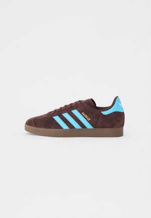 Brązowe zamszowe sneakersy z niebieskim wzorem trzech pasków i złotym napisem "Gazelle". Posiadają teksturowaną brązową gumową podeszwę oraz wyściełany język.