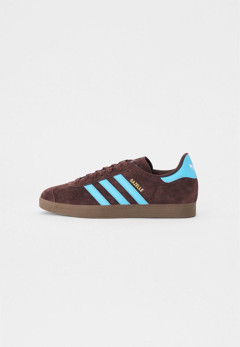 Brun mockasinsneaker med blå tre-randers design och guld "Gazelle" text. Har en texturerad brun gummisula och vadderad tunga.