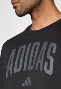 Svart bomulls-t-skjorte med en stor grå "ADIDAS"-logo på forsiden, med rund hals og korte ermer, teksturert stoff.