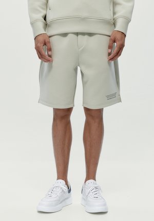 Hombre con pantalones cortos grises claros hasta la rodilla y sudadera a juego, zapatillas blancas, de pie contra un fondo liso.